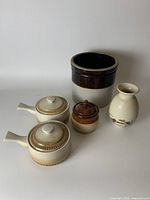 Photo showing full assortment of five pottery items on a white background: dark brown and off-white crock, porcelain vase, two ceramic soup bowls with lids and handles, and small brown jar.