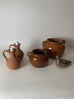 Five assorted pottery pieces displayed: two large brown glazed bean crocks, two small terracotta jugs with white lids, and a bird figurine.