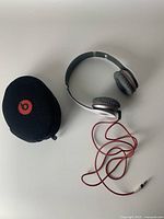 Top view of Beats by Dr Dre headphones with red cable coiled beside black soft carrying case featuring red Beats logo.