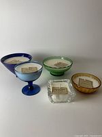 Full view of 5 handmade soy wax candles in varied vintage vessels, showing different colors and styles of holders with visible labels.