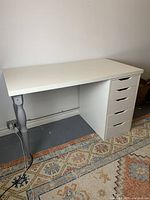 Full front and side view of the white IKEA desk showing grey legs and ALEX drawers on a patterned rug.