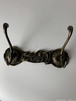 Front side view of both antique Italian brass wall hooks connected on a decorative backplate showing floral and wing motifs, with curved hooks.