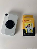 Kodak Printomatic instant camera with lens, flash, and Kodak logo, next to unopened box of Kodak ZINK photo paper with yellow packaging.