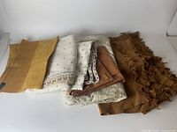 Folded assorted vintage table linens including brown fringed sets, white lace embroidered cloths, and mustard colored woven cloth with tag visible.