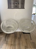 Pair of white-painted rattan hoop chairs with black metal legs, arranged side by side on a wooden floor in front of a white wall with decorative wooden panel above.