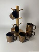 Group photo showing six mid century modern ceramic coffee mugs with brown and tan leaf-like pattern placed on and around a wooden mug stand with six pegs.