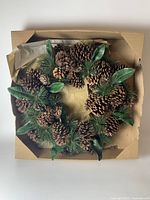 Full overview of the pinecone wreath in a cardboard box showing large pinecones, green pine needles, and green leaves