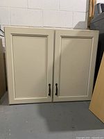 Front view of cream-colored storage cupboard with two paneled doors and dark handles on a gray concrete floor.