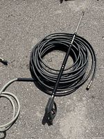 Coiled black high pressure washer hose with attached long spray gun and additional gray hose beside it lying on asphalt.