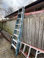 Werner 8 foot extension ladder leaning against a wooden fence outdoors, showing blue sides and aluminum steps with paint marks.