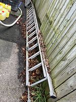 Full length side view of aluminum extension ladder showing metal construction, paint spots, and dried leaves in between rungs