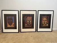 Three framed art prints displayed side by side on floor, each with stylized abstract face and signed and numbered by artist.
