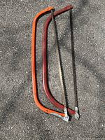 Two large bow saws with orange and red metal frames and attached serrated blades placed on asphalt surface
