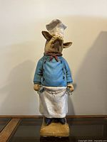 Front view of decorative ceramic pig chef figurine wearing blue jacket, white apron, red neckerchief, and chef's hat, standing on tan base.