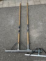Two floor squeegees with wooden handles laid on pavement, showing front side with metal blades and rubber strips in good condition.