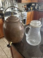 Photo shows the solid hammered copper drink dispenser with curved brass handle, lid, and spout along with the translucent plastic jug with lid and handle, both placed on a kitchen counter.