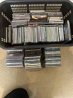 Large plastic basket filled with multiple music CDs in jewel cases, with three stacks of CDs laid on the floor next to it.
