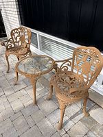 Side view of two ornate cast iron outdoor chairs and round glass-top table with rose motif, placed on a patio.