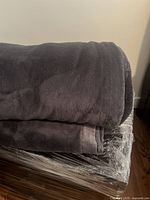 Rolled and folded thick fuzzy blanket in dark gray/purple tone shown lying on wrapped base.