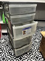 Full view of the plastic storage bin showing 5 transparent drawers in black frame with one drawer partially open on tiled floor.