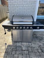 Front view of stainless steel propane BBQ with six knobs, closed cabinet doors, and side burner on black frame.