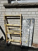 Yellow rolling baker scaffolding frame standing on paved ground against a brick wall, showing paint splatters and wear.