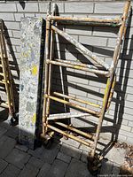 Photo shows used metal scaffolding frame with diagonal bracing and caster wheels, alongside a wooden platform heavily coated with multiple layers of dried paint.