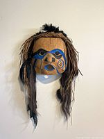 Front view showing full carved wooden mask with painted facial patterns, braided grass and feather details.