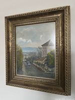 Framed vintage landscape painting showing countryside road with horse-drawn carriage and building viewed from the side, highlighting the ornate gold frame.