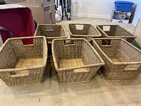 Seven storage bins arranged on carpet floor: six woven natural-tone bins with cut-out handles and one cloth bin with black-and-white paisley fabric outside and fuchsia pink lining inside visible in background.