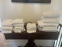 Multiple stacks of various white towels, including bath towels, hand towels, and washcloths, arranged on a wooden table.