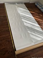 Writable magnetic wall covering rolled out on hardwood floor with measuring tape showing a width of 48 inches and length 96 inches (approx). Light shadows fall on the white surface revealing slight indentations.