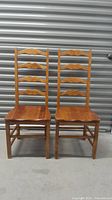 Pair of vintage hardwood dining chairs with ladder backs and flat wood seats placed side by side on concrete floor in front of a metal storage door.