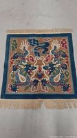 Full view of the small square Japanese vintage rug on a flat surface showing the whole dragon motif, color palette, and tassels.