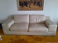 Front view of a large white leather sofa with two seats and cushioned backrest, sitting on a mustard carpet, with a painting on the wall behind.