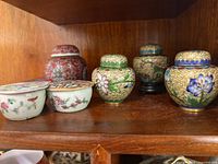 Image showing a group of cloisonné style spice jars and small ceramic lidded cups placed on a wooden shelf, clearly depicting intricate enamel floral designs and hand-painted scenes.