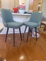 Photo showing front and side view of two blue upholstered swivel bar stools with wooden legs and chrome footrests, positioned on hardwood floor.