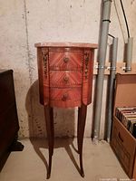 Full front view of the tall wooden cabinet showing stone top, three drawers with wood inlay and brass knobs, and slender legs.