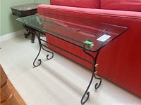 Side angle photo showing the full length of the table with rectangular glass top and black metal frame in front of a red couch.