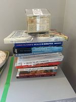 Stack of assorted cookbooks with plastic container of recipe cards on top.