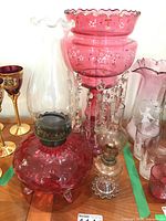 Overall display showing hobnail cranberry oil lamp foreground, clear oil lamp beside it, art glass vase in background, ruby stemware partially visible.