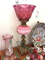 Full view of cranberry glass lamp with brass base, showing cranberry ruffled glass shade, pink decorative glass midsection, and ornate brass stand with figure decoration. Lamp is surrounded by other glassware and porcelain.