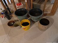 Photo showing two large green plastic plant pots, three smaller plastic pots including a yellow bucket, and a tan stoneware crock on concrete floor