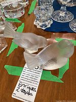 Two frosted glass dove figurines resting on a table among other glassware. The doves have textured feathers and a matte finish.