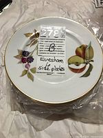 Top view of a single side plate showing the Evesham fruit and leaf design with gold rim and a sticker label noting the lot details.