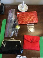 Three coin purses (black leather-like, red fabric, orange with gold pattern), ceramic ring dish with floral design, assorted metal trinkets in plastic bag on wooden surface