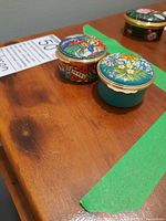 Two enamel jewelry boxes on a wooden surface showing colorful floral and stained glass designs with gold-tone edging