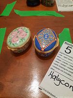 Photo showing two Halcyon Days enamel jewelry boxes, one with pink rose design and the other with blue and gold ornate pattern on a wooden surface.