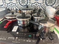 View of 4 stainless steel pots with lids and black handles, plus 2 red non-stick frying pans without lids, arranged on a KitchenAid stove.