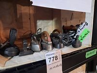 Seven miniature antique irons arranged in a row inside a white box, showing various handle types and sizes.